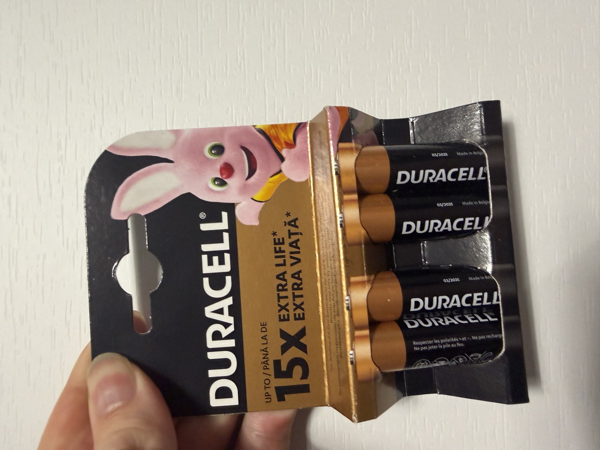 A hand holding a pack of four Duracell AA batteries, the pink Duracell bunny peeking from the corner